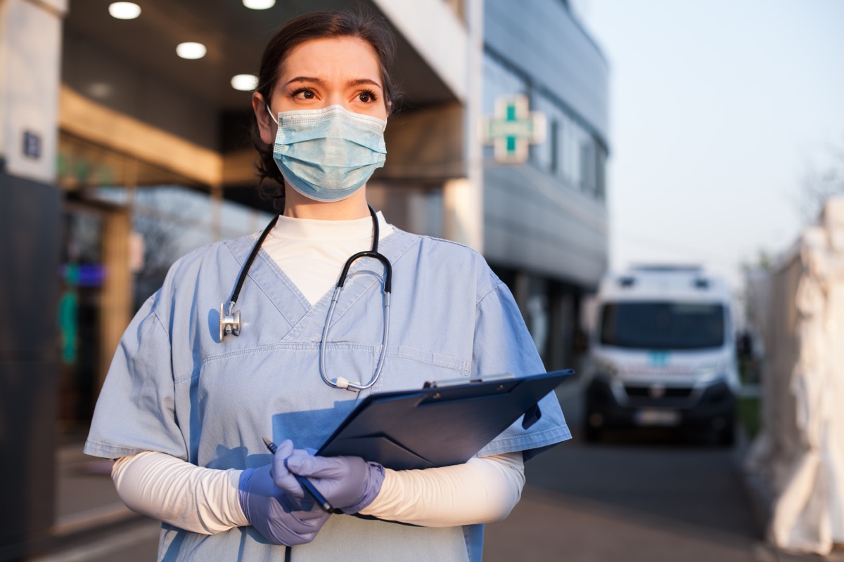 Young female EMS key worker doctor in front of healthcare ICU facility, wearing protective PPE face mask equipment,holding medical lab patient health check form