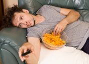 Lazy man in a gray t-shirt laying on the couch with the remote and a bowl of potato chips.