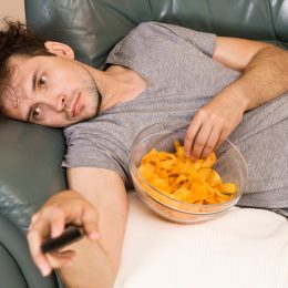 Lazy man in a gray t-shirt laying on the couch with the remote and a bowl of potato chips.