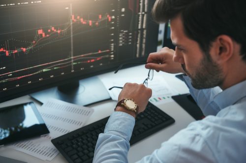 Bearded male trader looking at watch on his hand while working with data and charts on computer screens in his modern office.
