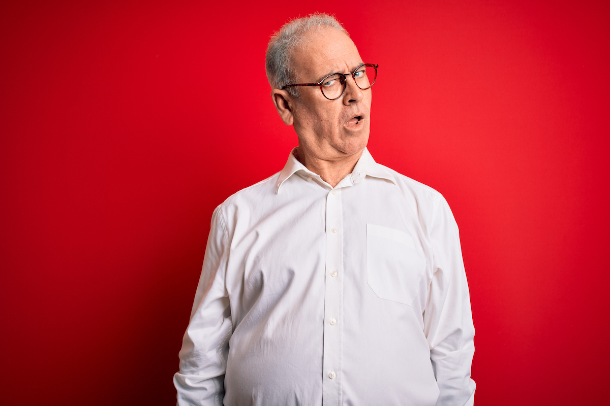 A middle-aged gray-haired man wearing a white button-down shirt and glasses over a red background, looking skeptical and sarcastic, surprised with open mouth