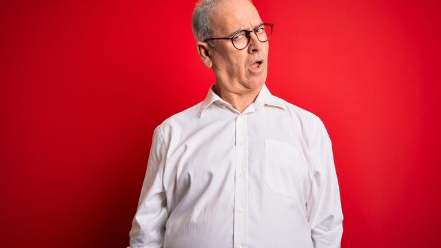 A middle-aged gray-haired man wearing a white button-down shirt and glasses over a red background, looking skeptical and sarcastic, surprised with open mouth
