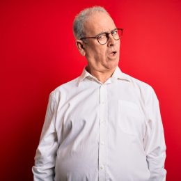 A middle-aged gray-haired man wearing a white button-down shirt and glasses over a red background, looking skeptical and sarcastic, surprised with open mouth