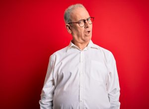 A middle-aged gray-haired man wearing a white button-down shirt and glasses over a red background, looking skeptical and sarcastic, surprised with open mouth