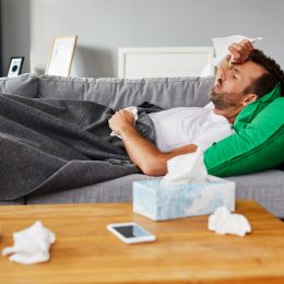 A man lying on the couch with tissues around him feeling symptoms of COVID-19 or the flu