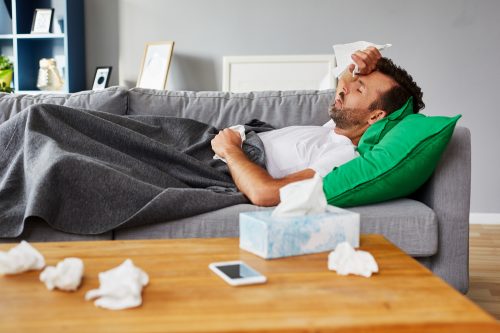 A man lying on the couch with tissues around him feeling symptoms of COVID-19 or the flu