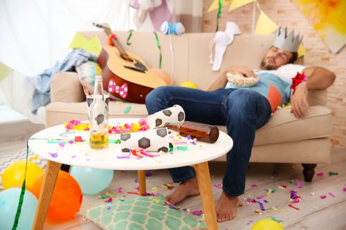A drunk man asleep on the couch after a party, with a mess surrounding him.