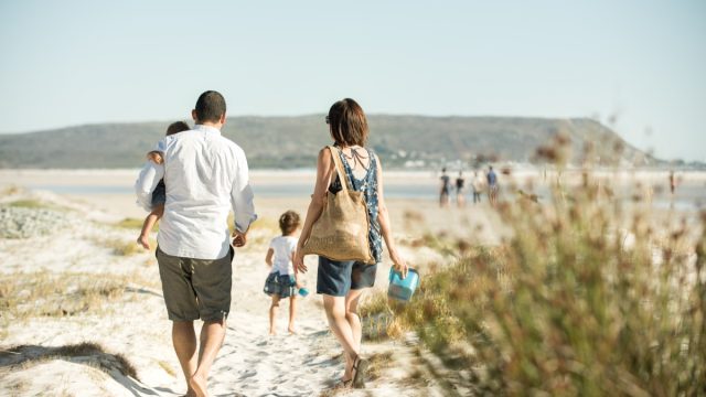 family walking on the beach outdoors in the sun, carrying their son while their daughter leads the way
