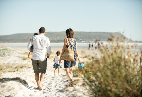 οικογένεια που περπατά στην παραλία υπαίθρια στον ήλιο, κουβαλώντας τον γιο της ενώ η κόρη τους οδηγεί το δρόμο