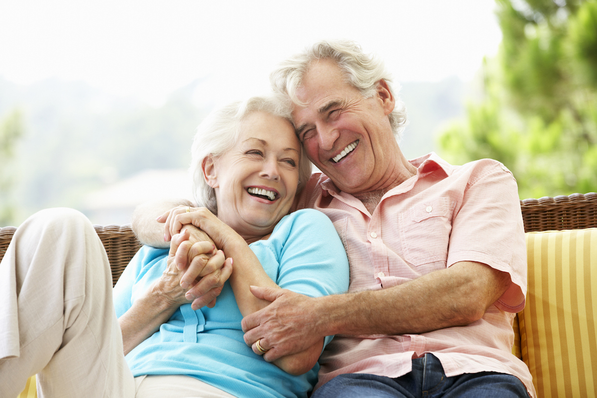 An older couple, both with white hair, sitting on a bench, hugging and laughing.