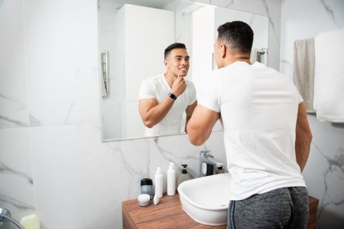 Morning hygiene concept. Back side waist up portrait of young brunette muscular smiling guy in white t-shirt looking at reflection on mirror and touching his chin