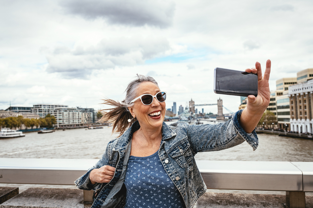 A senior woman taking a selfie in London with the Tower Bridge behind her.