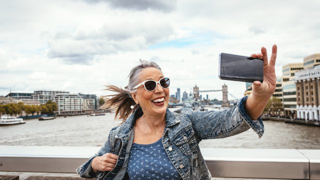 A senior woman taking a selfie in London with the Tower Bridge behind her.