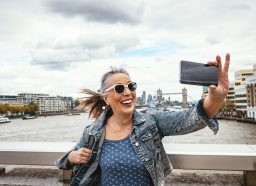 A senior woman taking a selfie in London with the Tower Bridge behind her.