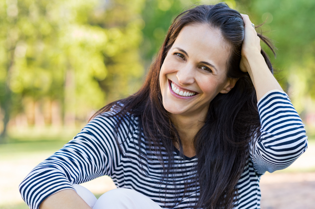 A smiling woman in a park holding back her dark hair.