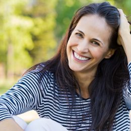 A smiling woman in a park holding back her dark hair.