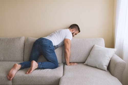 Bearded man on knees searching behind sofa lost thing.