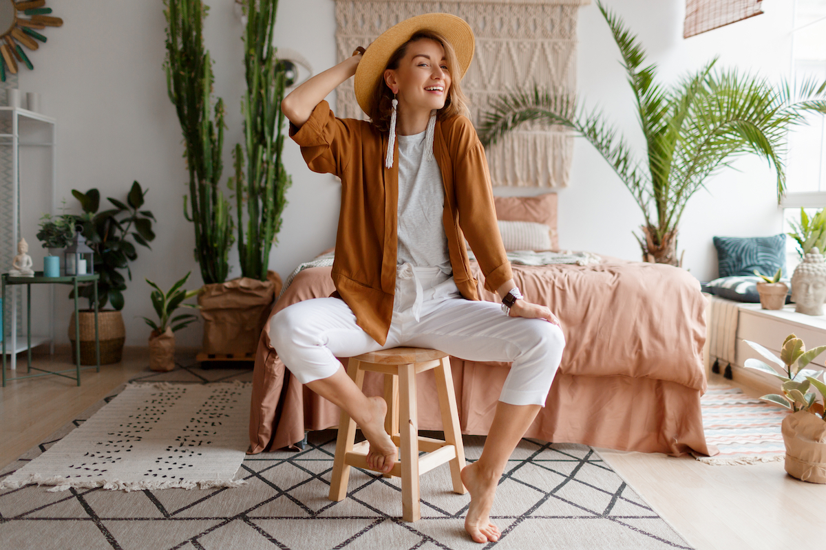 A stylish young woman wearing linen clothes sitting on a stool in front of her bed in a bohemian-style room.