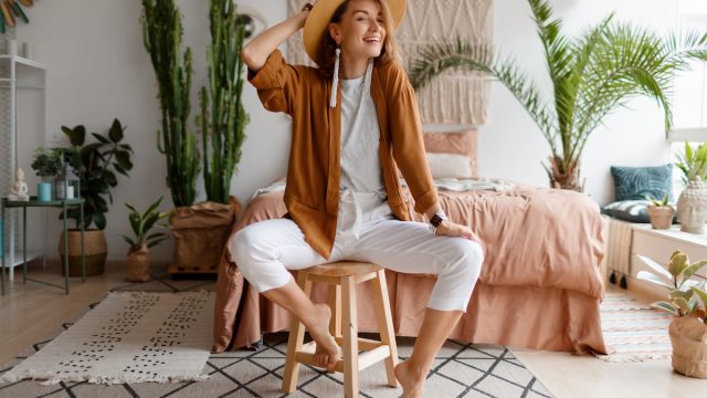 A stylish young woman wearing linen clothes sitting on a stool in front of her bed in a bohemian-style room.