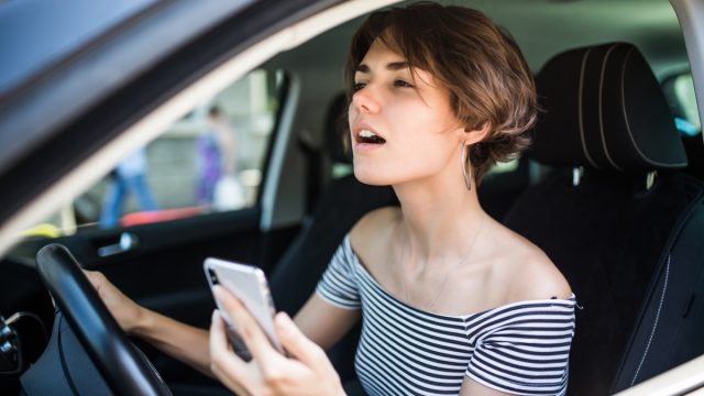 Closeup portrait, young woman driving in black car and checking her phone, annoyed by navigation gps system or bad text message or email, isolated outdoors background