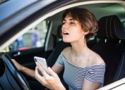 Closeup portrait, young woman driving in black car and checking her phone, annoyed by navigation gps system or bad text message or email, isolated outdoors background
