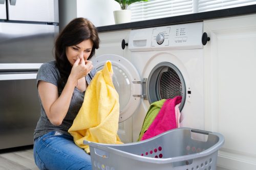 Young Woman Looking At Smelly Clothes Out Of Washing Machine In Kitchen