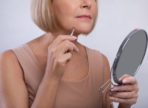 Cropped view of mature woman removing unwanted hair from her chin, using tweezers, looking in mirror