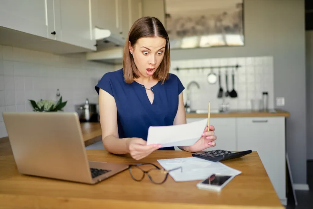 Surprised young woman using a laptop computer sitting at her kitchen holding utility bill and bank statements.