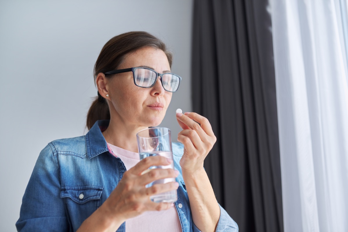Mature middle-aged woman in casual clothes at home holding pill and glass of fresh water