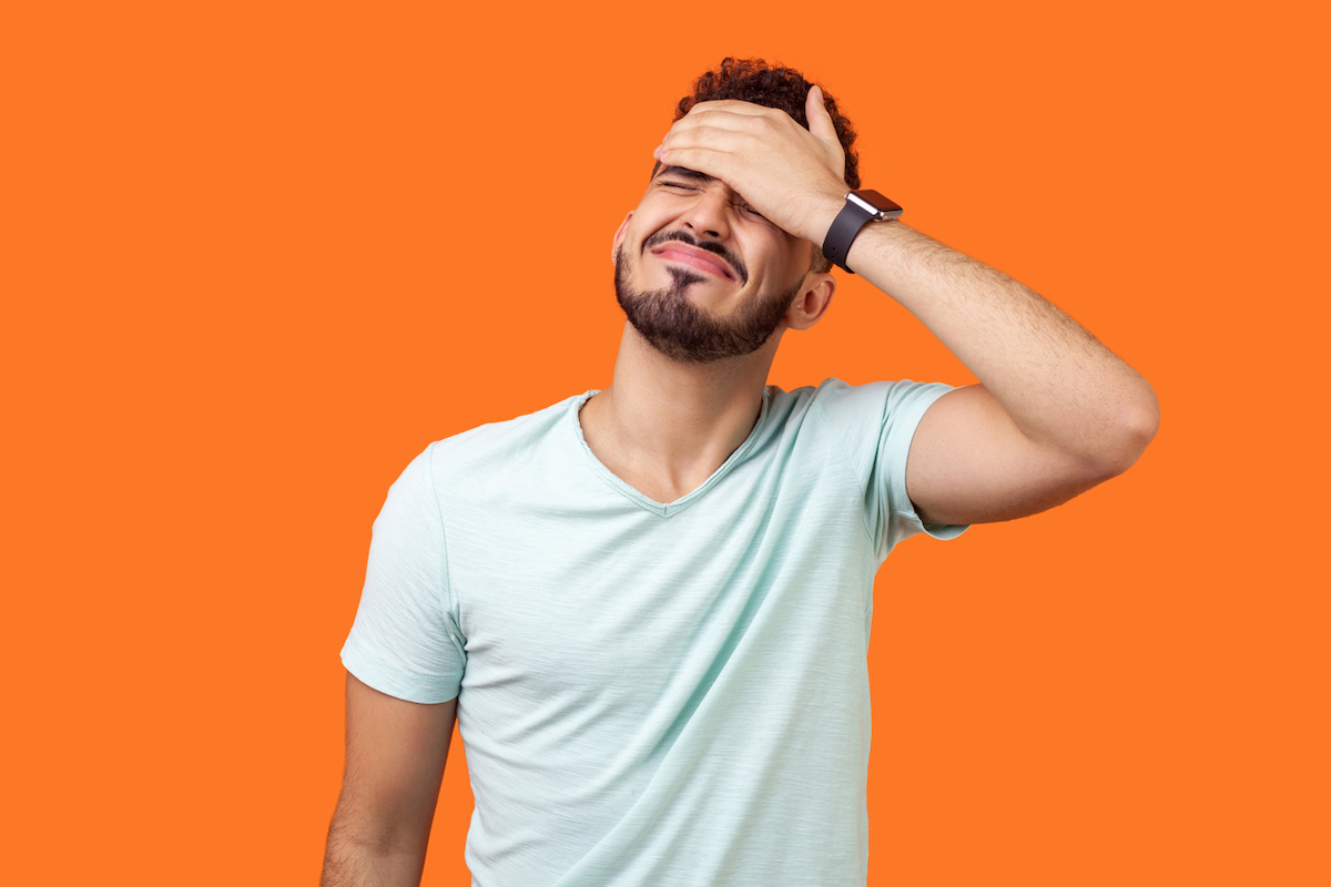 Young man with a beard and white t-shirt against an orange background putting his palm to his face in embarrassment