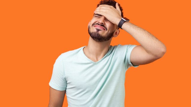 Young man with a beard and white t-shirt against an orange background putting his palm to his face in embarrassment