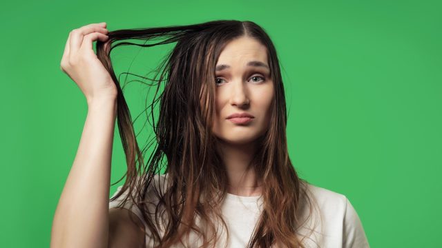 A young woman against a green background looking upset about her oily hair.