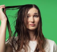 A young woman against a green background looking upset about her oily hair.