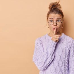 Young woman presses index finger over lips looks mysteriously away