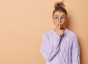 Young woman presses index finger over lips looks mysteriously away