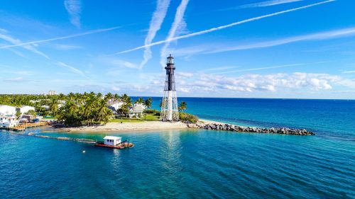 The Hillsboro Inlet Lighthouse in Fort Lauderdale, Florida. The black and white structure is set on a sandy shore with the teal-colored ocean.