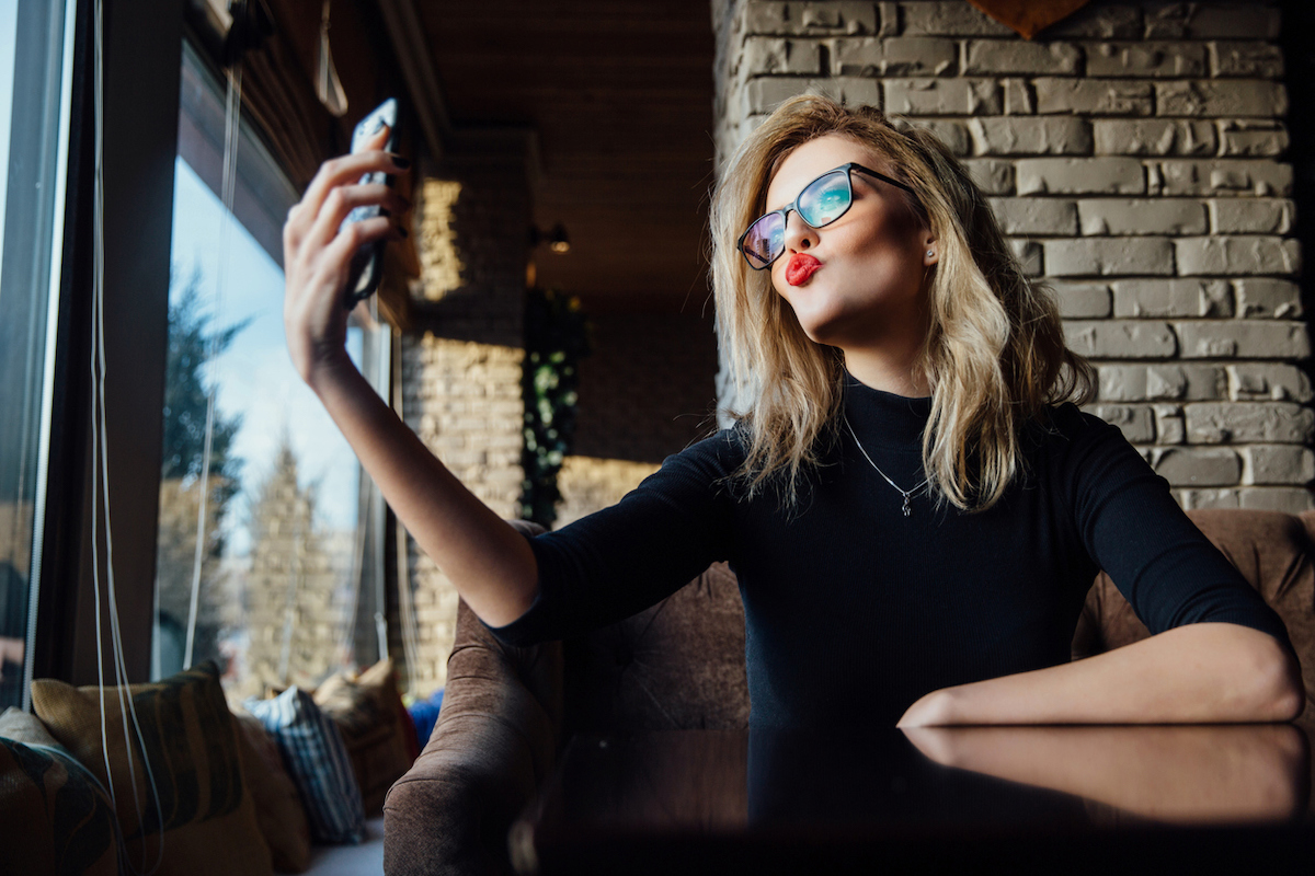 Blonde woman wearing a black turtleneck, red lipstick, and glasses taking a selfie in coffee shop