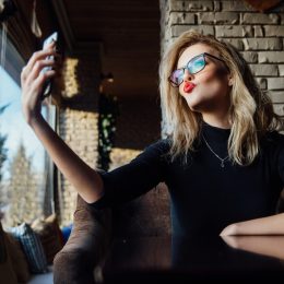 Young beautiful blonde woman taking a selfie in coffee shop. Hipster, red lips, glasses. Against the background of a brick stylish design wall.