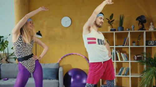 A young couple dressed up as 1980s aerobics instructors doing a workout in their living room.