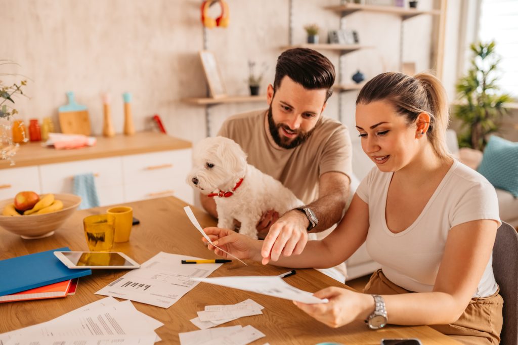 A young couple sitting at the table checking their finances, while the man holds a small white dog.