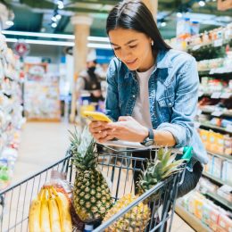 Happy young woman shopping for groceries in supermarket standing near her trolley using smartphone