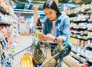 Happy young woman shopping for groceries in supermarket standing near her trolley using smartphone