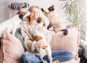 A happy blonde woman on her couch playing with her two dogs and cat.
