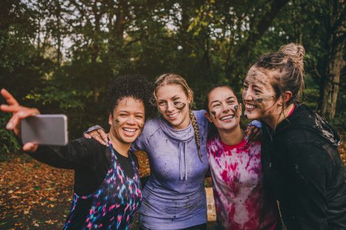 Four women are taking a selfie together on a smart phone after completing a charity obstacle course. They are wearing sports clothing and are covered in mud and water.