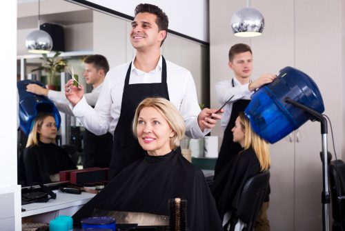 Male stylist cutting hair of older blonde woman in salon.