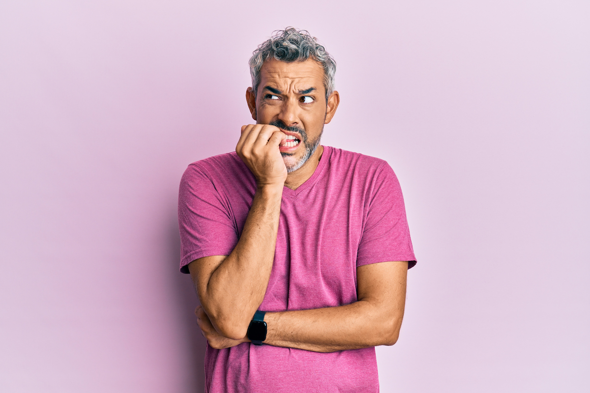 A middle-aged man with gray hair and a purple t-shirt nervously biting his nails looking insecure against a light purple background