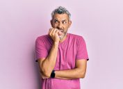 A middle-aged man with gray hair and a purple t-shirt nervously biting his nails looking insecure against a light purple background