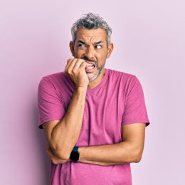 A middle-aged man with gray hair and a purple t-shirt nervously biting his nails looking insecure against a light purple background