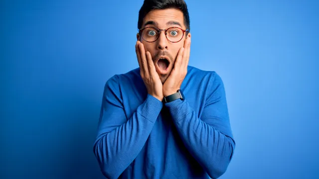 Young handsome man with beard wearing casual blue sweater and glasses over blue background afraid and shocked