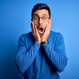 Young handsome man with beard wearing casual blue sweater and glasses over blue background afraid and shocked